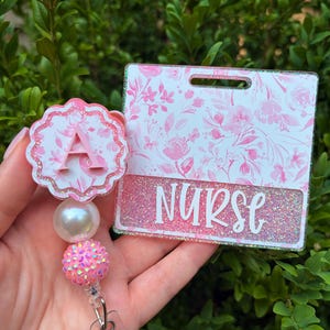 May include: A badge reel and matching ID holder set featuring a pink floral pattern. The circular reel has a scalloped edge with a letter A, a white pearl bead, and a pink rhinestone bead. The rectangular ID holder displays the word NURSE in white.