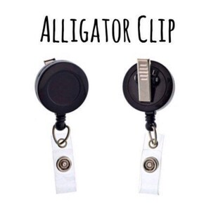 Pu&ograve; includere: Porta badge retrattile nero con clip a coccodrillo e cinturino in plastica trasparente. Il testo "ALLIGATOR CLIP" &egrave; stampato su sfondo bianco.