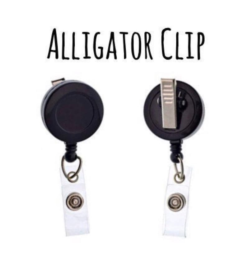 Pu&ograve; includere: Porta badge retrattile nero con clip a coccodrillo e cinturino in plastica trasparente. Il testo "ALLIGATOR CLIP" &egrave; stampato su sfondo bianco.