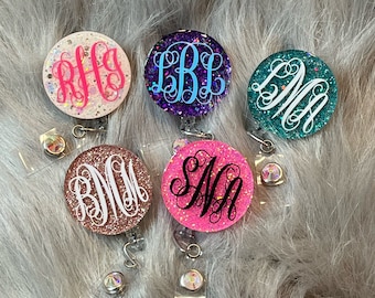 May include: Five glitter badge reels with different monogram initials. The reels are pink, white, purple, teal, and gold. Each reel has a clear plastic clip and a silver swivel.