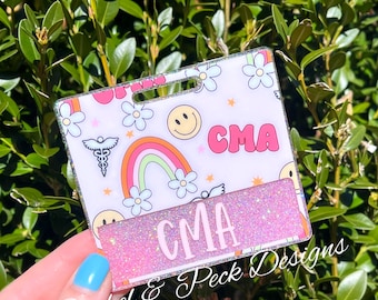May include: A pink and white glitter badge with a rainbow, flowers, and a smiley face. The badge says "CMA" in pink letters and "Bushel & Peck Designs" in black letters.