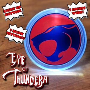 May include: A round, light-up button with a blue and red graphic of a cat-like creature. The button is 2 inches wide and has 3 blink settings. Text on the button reads "Eye of Thundera" and "Light Up Button!"
