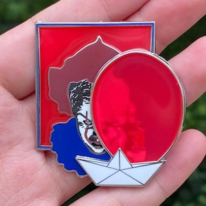 May include: Enamel pin featuring a red balloon, a paper boat, and a portrait of a clown with white face paint, red hair, and a blue collar. The pin has a red, white, blue, and brown color scheme.