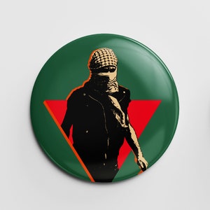 May include: A circular badge with a green background and a red triangle. The design features a figure in black clothing and a patterned head covering. The badge has a glossy finish.