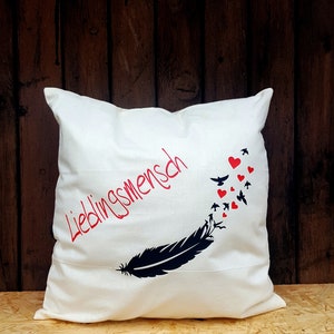 Könnte beinhalten: Ein weißes Kissen mit einem schwarzen Feder-Design und roten Herzen. Der Text "Lieblingsmensch" ist in Rot auf dem Kissen aufgedruckt.