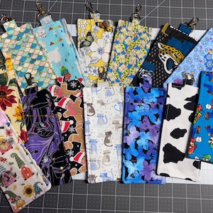 May include: A collection of fabric eyeglass cases with various patterns, including floral, animal print, and cartoon characters. Each case has a metal clip for attachment. The cases are in various colors, such as blue, yellow, and black.