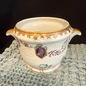 May include: A white porcelain bowl with gold trim and handles. The bowl features a decorative crest and floral patterns in various colors. The rim and base have gold accents, and the bowl sits on a small foot.