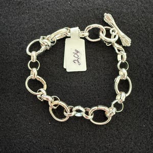 May include: A silver bracelet with a toggle clasp and a series of interconnected oval links. The toggle clasp is adorned with small, sparkling embellishments. A white tag with the number "206" is attached to the bracelet.