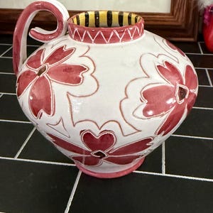 May include: A white ceramic pitcher featuring a hand-painted pattern of large, dark pink flowers with embossed outlines. The pitcher has a curved handle, a pink base, and a rim decorated with alternating yellow and black vertical stripes.