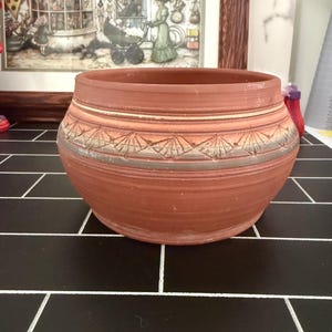 May include: A terracotta bowl with a wide, rounded body and a smooth, open top. The exterior features a horizontal band of carved, fan-shaped patterns encircling the middle, accented with a subtle grey wash over the natural reddish-brown clay.