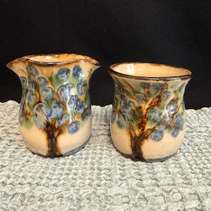 May include: Two ceramic vases with a tree design. The vases have a beige base with brown tree trunks and blue and green foliage. The rims are a darker brown. The vases are on a gray textured surface.