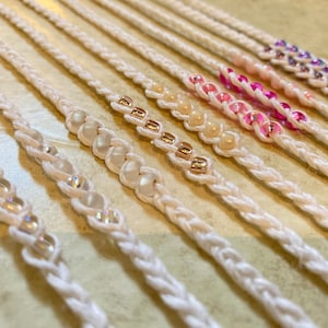 May include: A set of white braided bracelets with different colored beads. The beads are in a rainbow pattern, starting with clear beads, then white, gold, pink, and purple.
