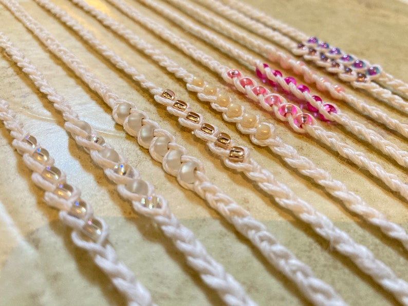 May include: A set of white braided bracelets with different colored beads. The beads are in a rainbow pattern, starting with clear beads, then white, gold, pink, and purple.