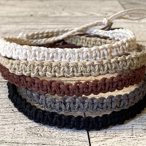 May include: Five braided bracelets in different colors, including white, beige, brown, gray, and black. The bracelets are stacked on top of each other, with the white bracelet on top.