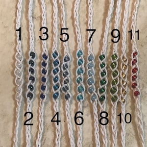 May include: Eleven white cord bracelets with different colored beads. The bracelets are numbered 1 through 11.
