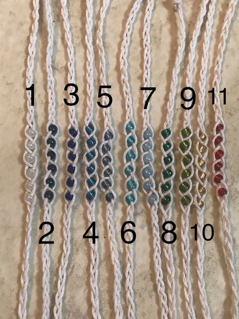 May include: Eleven white cord bracelets with different colored beads. The bracelets are numbered 1 through 11.