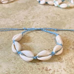 May include: A light blue cord bracelet featuring six white cowrie shells strung along the center. The bracelet has adjustable knotted ends. Two additional bracelets with cowrie shells are visible in the background, one with tan cord and one with teal.