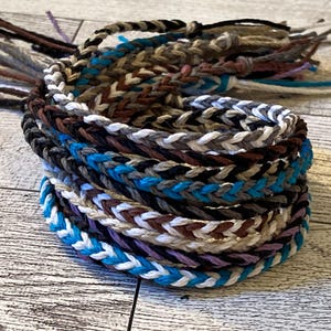 May include: A stack of braided bracelets in various colors, including white, brown, black, blue, and purple. The bracelets are made of a woven cord material and are arranged in a circular pattern.