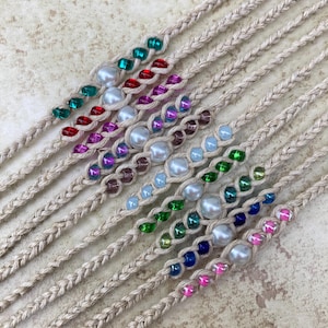 May include: A collection of braided beige cord bracelets, each featuring a central row of alternating white faux pearls and colorful translucent beads in shades of red, green, blue, purple, pink, and light blue, arranged in a parallel diagonal pattern.