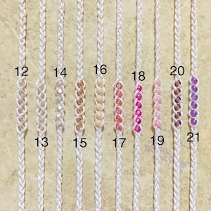 May include: A close-up of ten white macrame cords with beads. The cords are numbered 12 through 21. The beads are different colors, including white, gold, pink, and purple.