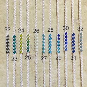 May include: Ten white cords with different colored beads strung on them. The cords are numbered 22 through 32. The beads are in various shades of blue, green, and white.