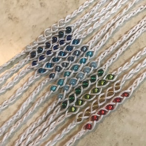 May include: A set of handmade bracelets with a rainbow of colored beads woven into white cord.