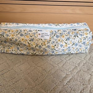 Peut inclure: Une trousse de maquillage rectangulaire à imprimé floral avec une fermeture éclair bleu clair et une petite poignée sur le côté. Le sac présente un fond blanc avec des fleurs bleues et jaunes. Le texte "With love, The Sunday Nest" est sur une petite étiquette blanche.