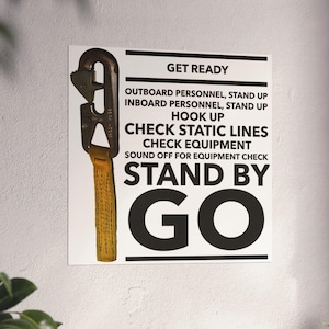 Könnte beinhalten: Ein weißes Poster mit schwarzem Text, das Anweisungen zur Ausrüstung enthält, darunter "GET READY" und "STAND BY GO". Ein Metallwerkzeug mit gelbem Gurt ist am Poster befestigt. Das Poster hängt an einer Wand.