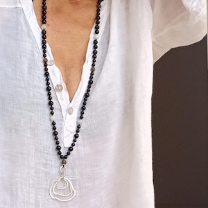 Könnte beinhalten: Eine lange schwarze Perlenkette mit einem silbernen Anhänger in Form eines Kreises mit einem kleineren Kreis darin. Die Kette wird von einer Person getragen, die ein weißes Hemd trägt.