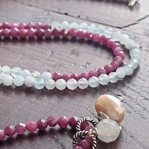 May include: Close-up of three beaded necklaces. The top two necklaces feature light blue beads and the bottom necklace has deep red beads. The bottom necklace has a pearl and a white stone pendant. The necklaces are on a wooden surface.