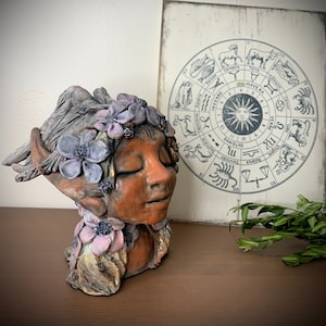 Puede incluir: Una escultura de cerámica de una cabeza de mujer con los ojos cerrados, que lleva una corona de flores. La escultura está pintada en tonos marrones, rosas y morados. El fondo presenta una rueda zodiacal vintage.