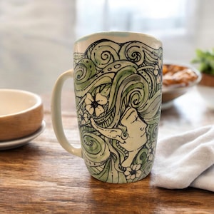Op de afbeelding: Een keramische mok met een handgeschilderd ontwerp van een vrouwengezicht met groene en zwarte swirls en witte bloemen.