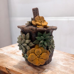 Peut inclure: Un récipient décoratif marron avec un couvercle, orné de succulentes sculptées dans des tons de vert et de jaune. Le récipient a un aspect texturé semblable à du bois et est placé sur une surface en bois.