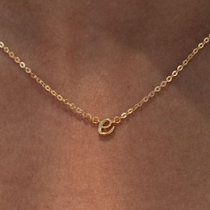 Puede incluir: Un delicado collar dorado con un pequeño colgante con la letra "e" en cursiva. La cadena es fina y el colgante está centrado, creando un aspecto minimalista y elegante. El collar está diseñado para ser usado cerca del cuello.