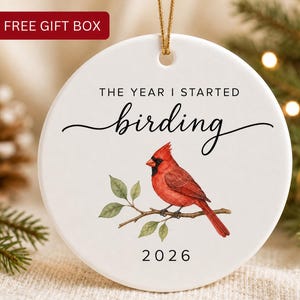May include: A round white ceramic ornament featuring a red cardinal bird perched on a branch. The text reads THE YEAR I STARTED birding and 2026. A gold string is attached to the top for hanging. The ornament has a smooth finish.
