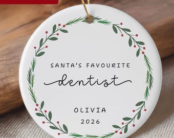 May include: A round white ceramic ornament featuring a green pine wreath with red berries. The text reads SANTA'S FAVOURITE dentist, OLIVIA, and 2026. The ornament hangs from a thin gold string against a neutral background.