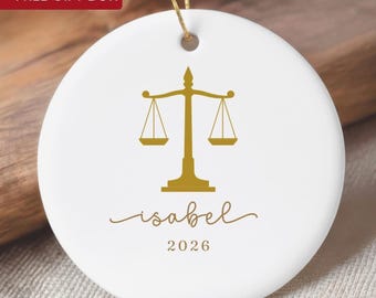 May include: A round white ceramic ornament featuring a gold scale of justice icon centered above the name Isabel in a cursive script and the year 2026 in a simple font. The ornament hangs from a thin gold string.