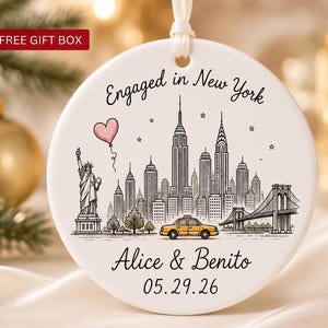 Puede incluir: Un adorno redondo de cerámica blanca con un dibujo de líneas negras del horizonte de Nueva York, la Estatua de la Libertad, un taxi amarillo y un globo de corazón rosa. El texto dice Engaged in New York, Alice & Benito y la fecha 05.29.26.