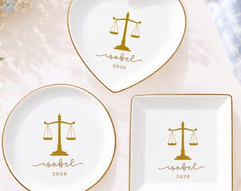 May include: Three white ceramic trinket dishes with gold rims, featuring a gold scale of justice icon, the name "isabel" in script, and the year "2026". The set includes one heart-shaped, one circular, and one square-shaped dish.