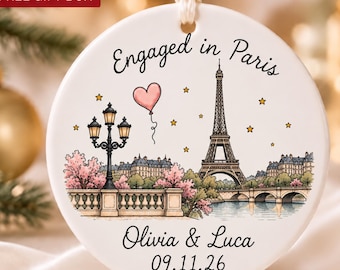 May include: A round white ceramic ornament featuring a printed illustration of the Eiffel Tower, a street lamp, and a pink heart-shaped balloon. The text reads Engaged in Paris, Olivia & Luca, and 09.11.26. It hangs from a white ribbon.