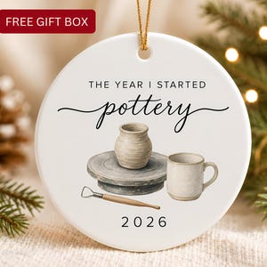 May include: A round white ceramic ornament featuring a gold hanging string. The design includes the text THE YEAR I STARTED pottery and 2026. An illustration shows a beige clay pot on a grey pottery wheel, a ceramic mug, and a metal sculpting tool.