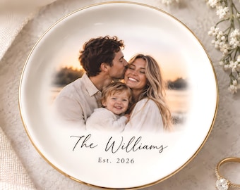 May include: A round white ceramic trinket dish with a gold-trimmed edge featuring a printed photograph of a family of three. The text "The Williams" is printed in black script above the text "Est. 2026" in a simple serif font.