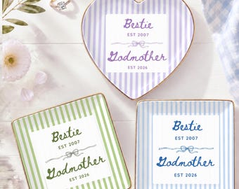 May include: A heart-shaped trinket dish with purple stripes and two square trinket dishes with green and blue stripes. Each dish features the text Bestie EST 2007, a bow graphic, and Godmother EST 2026. A gold ring sits near the heart dish.