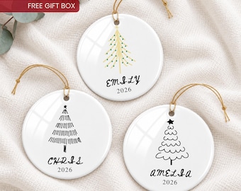 May include: Three round white ceramic ornaments with gold hanging strings. Each features a unique black or gold Christmas tree illustration, a personalized name in black script, and the year 2026 printed in black text below the name.