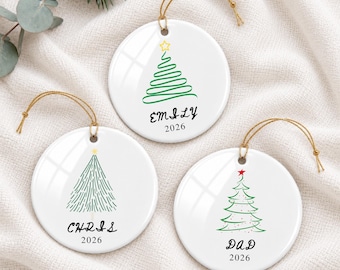 May include: Three round white ceramic ornaments with gold hanging strings. Each features a unique green Christmas tree illustration, a yellow or red star topper, and the printed names Emily, Chris, or Dad with the year 2026 below.