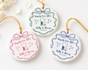 May include: Three round white ceramic ornaments with gold hanging strings. Each features a bow graphic, the text "Happily Ever After", "Kelly & Henry", and "10.05.26", plus a couple holding a heart. Colors are green, pink, and blue.