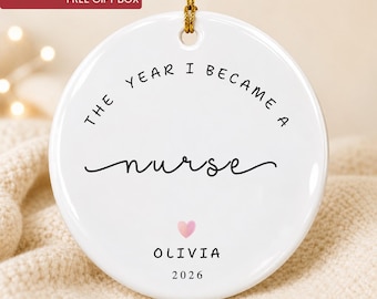 May include: A round white ceramic ornament featuring the black text THE YEAR I BECAME A nurse, a small pink heart, and the name OLIVIA followed by the year 2026. The ornament hangs from a gold string and has a smooth, glossy finish.