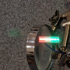 May include: A close-up of a small, rectangular LED display with red, white, and green lights. The display is mounted on a metal stand and is connected to wires.