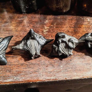 May include: Four dark grey water caltrops with a spiky design, arranged on a wooden surface. The caltrops have a textured, ridged appearance and are in a row. Blurred glass bottles are in the background.
