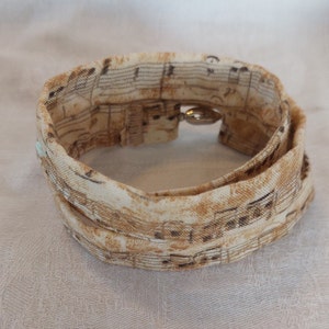 Könnte beinhalten: Ein weißes Stoffarmband mit braunem Noten-Druck und einem silbernen Verschluss.